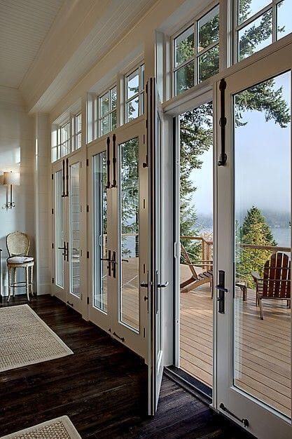 Large multi-panel French doors open to a spacious deck in a wooded setting, enhancing indoor-outdoor living with natural light and seating.