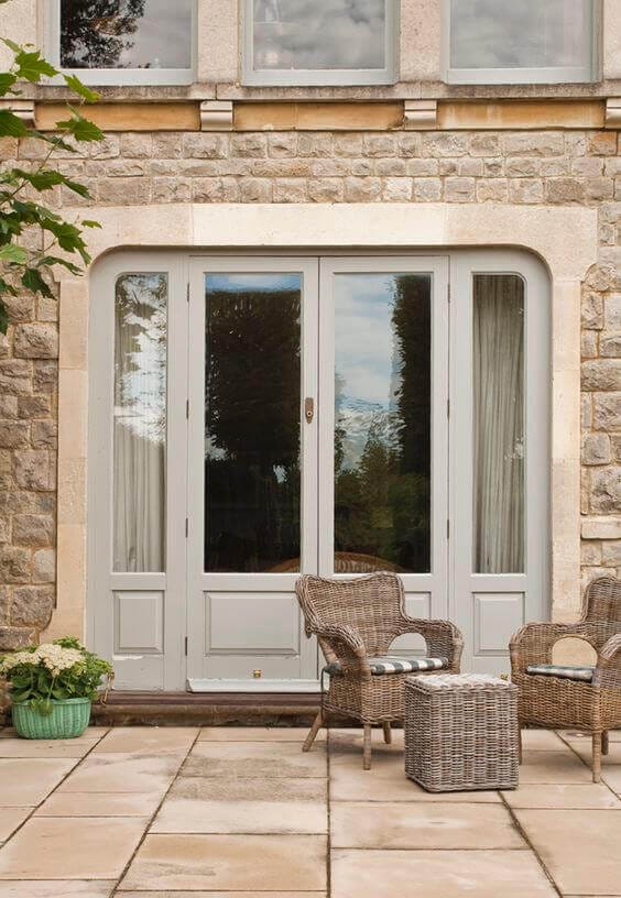 Double light gray French doors open to an outdoor patio with large stone tiles, surrounded by greenery and cozy seating.