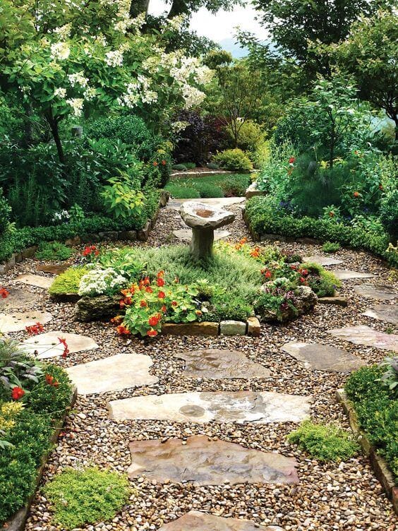 Flagstone and gravel walkway leads to a birdbath, bordered by lush greenery and vibrant flowers, enhancing the garden's tranquility.