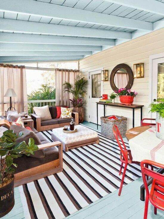 Cozy back porch design with a wooden sofa set and striped rug, surrounded by plants and curtains, perfect for outdoor living.