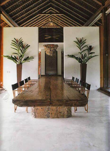 Long rustic live edge dining table, 3 meters long, surrounded by eight minimalist black chairs in a spacious, airy room with vaulted ceiling and green plants.