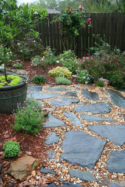 Natural flagstone and gravel walkway with vibrant flower beds, lush greenery, and a wooden fence, creating an organic, low-maintenance landscape.