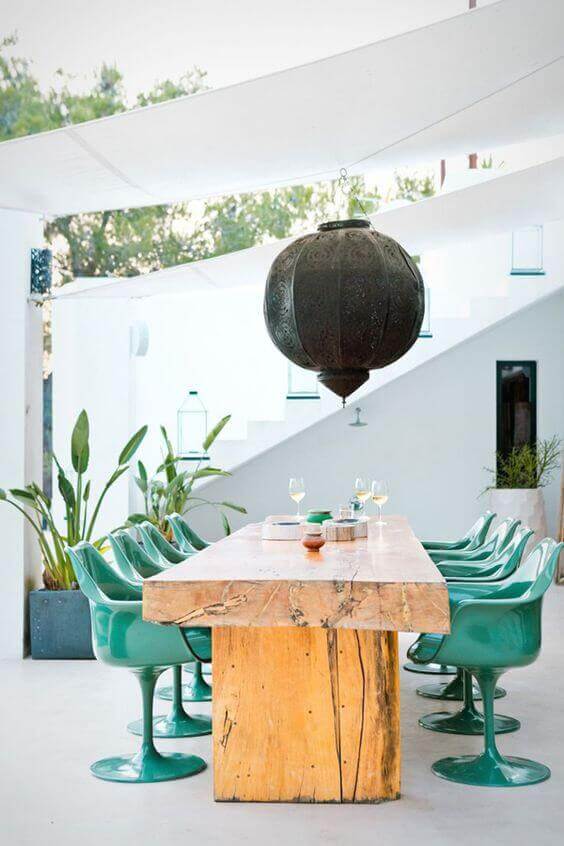 Long, rectangular live edge dining table in natural wood, surrounded by eight green molded plastic chairs, under a decorative pendant light.