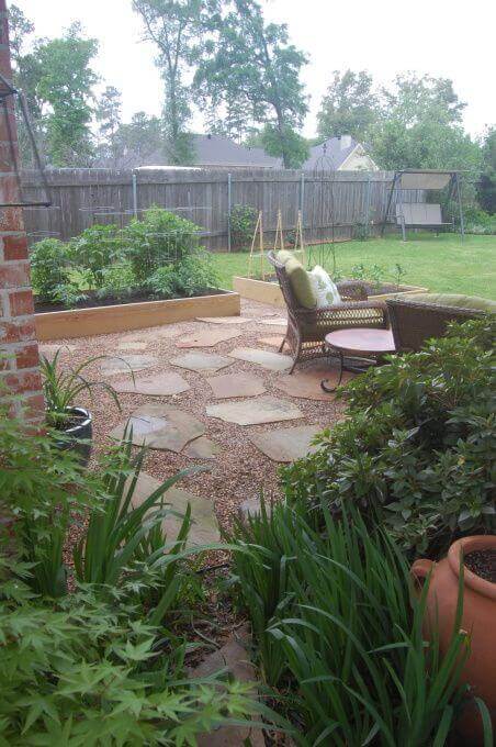 Flagstone and gravel walkway meanders through a lush garden, bordered by raised beds and a cozy seating area, creating a serene outdoor space.