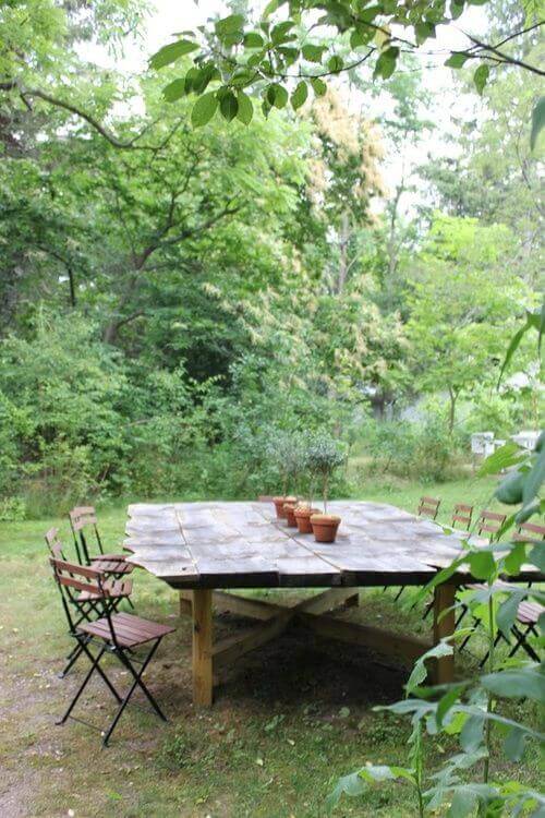 Large square live edge dining table made of reclaimed timber, surrounded by six folding chairs in a lush green outdoor setting.