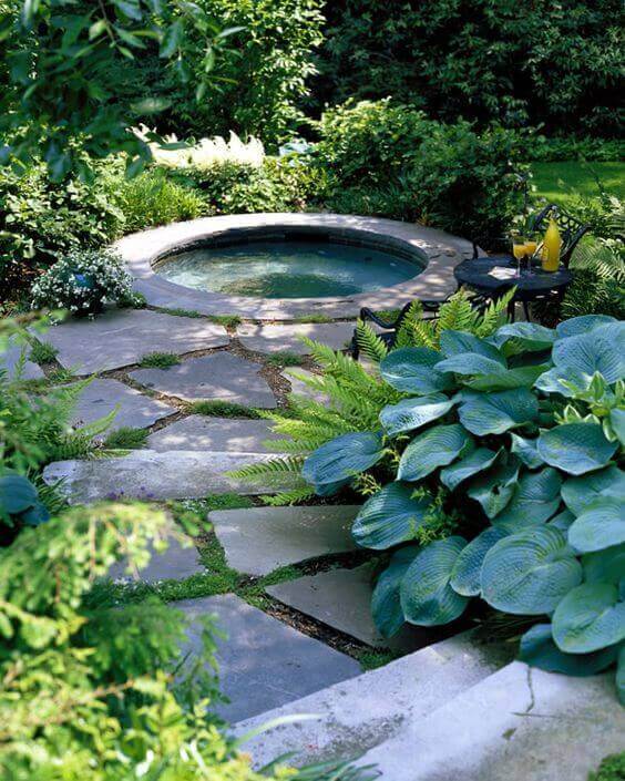 Circular swim spa surrounded by lush greenery and stone paths, featuring bluestone paving, low-growing plants, and wrought iron seating.