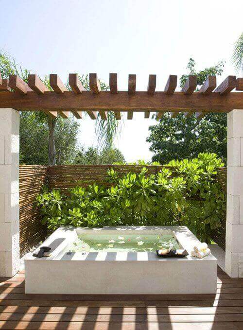 Rectangular outdoor swim spa, 2 meters long, with smooth stone sides and natural wood decking, surrounded by tropical plants and a wooden pergola.
