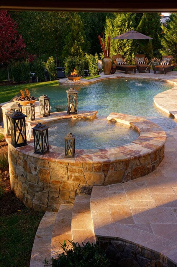 Swim spa with a circular, elevated spa on a stone wall, surrounded by paver stones and shaded lounge chairs, accented by decorative lanterns.
