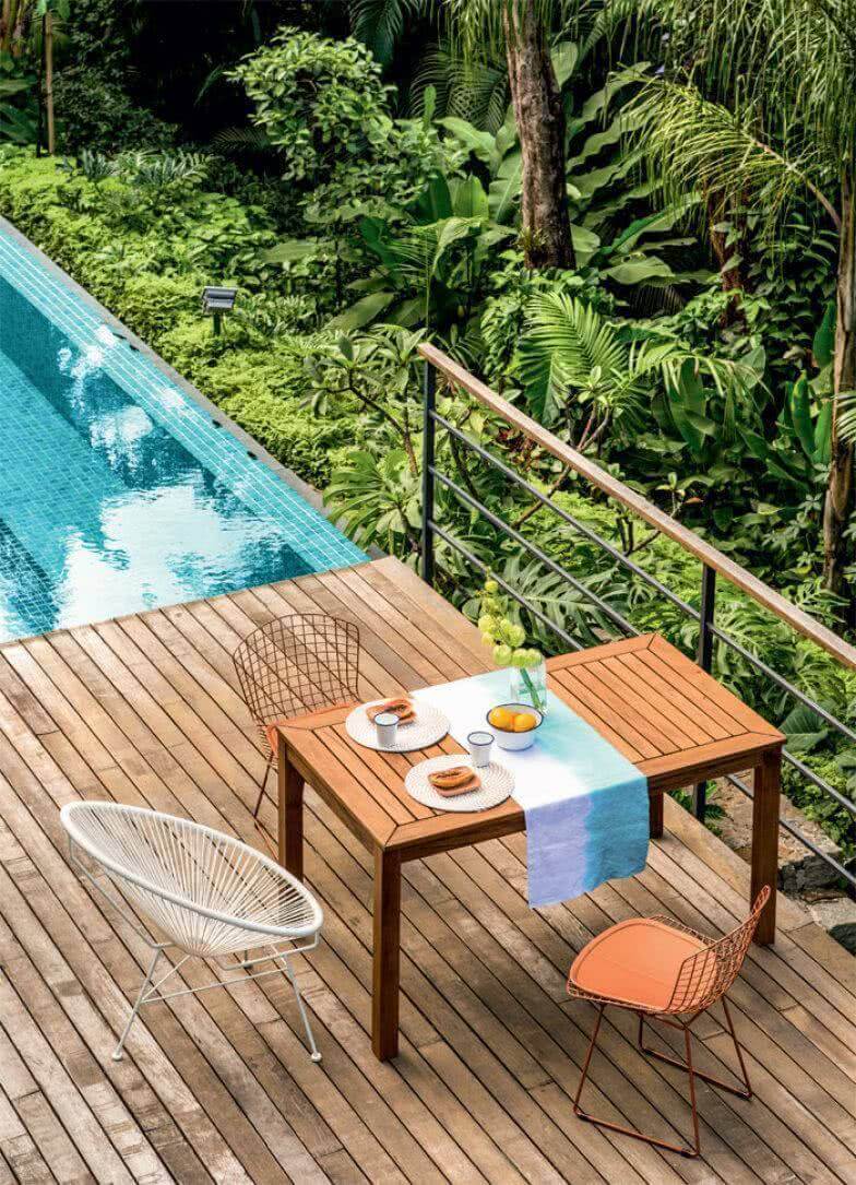 Outdoor dining area with a hardwood deck, featuring a wooden table, Acapulco chair, and a metal chair with an orange cushion.