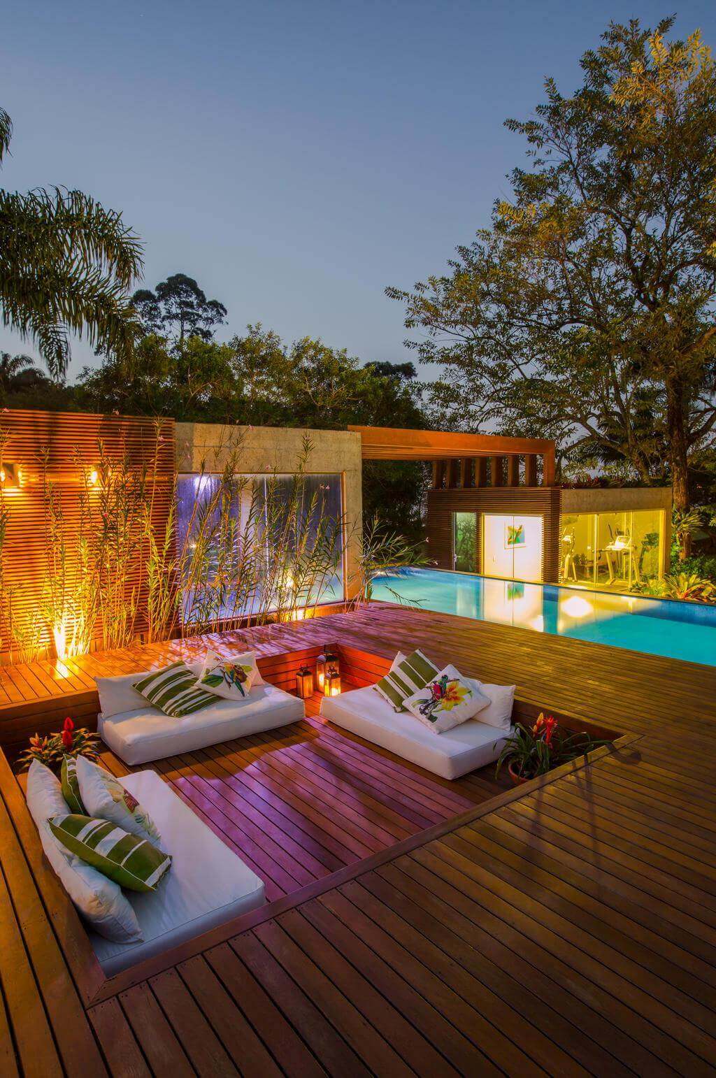 Wooden deck area with two white cushioned seating units, green-striped pillows, and ambient lighting. Surrounded by tropical plants and a pool.