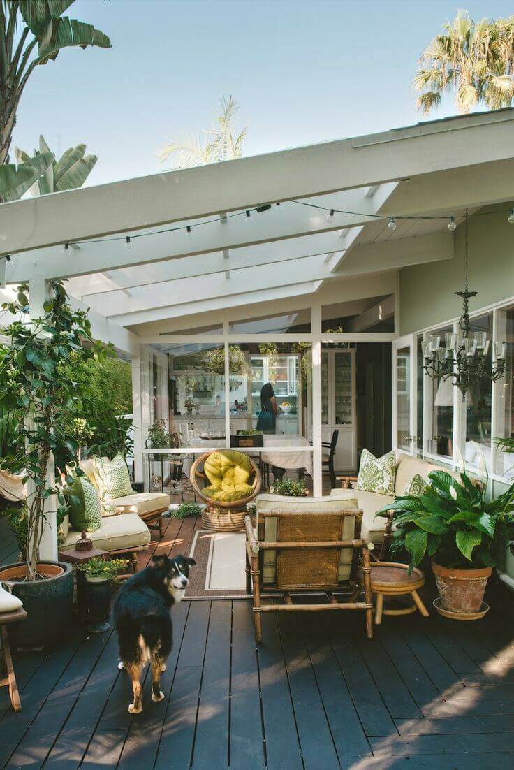 Outdoor living space with a covered wooden deck, featuring rattan seating, oversized cushions, and potted plants for a relaxing ambiance.