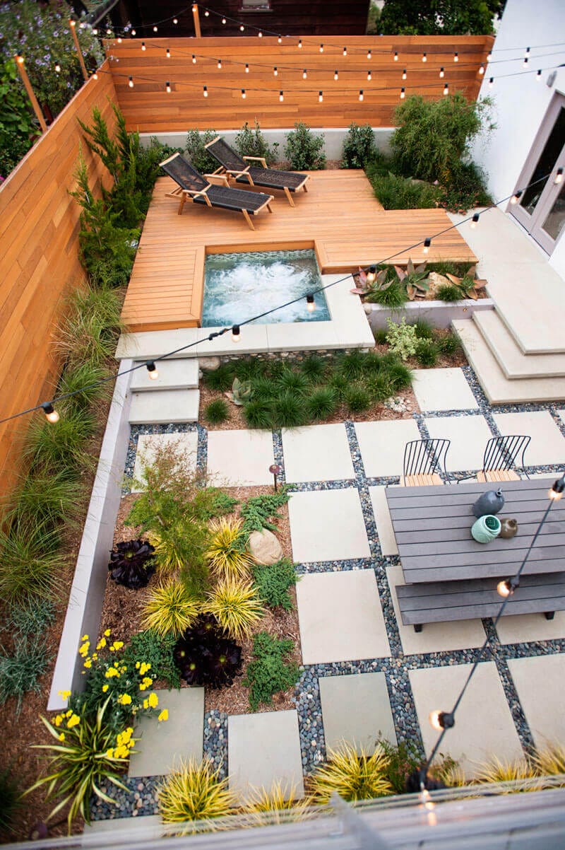 Compact backyard design featuring a wooden deck with lounge chairs, surrounded by concrete pavers and low-maintenance plantings.