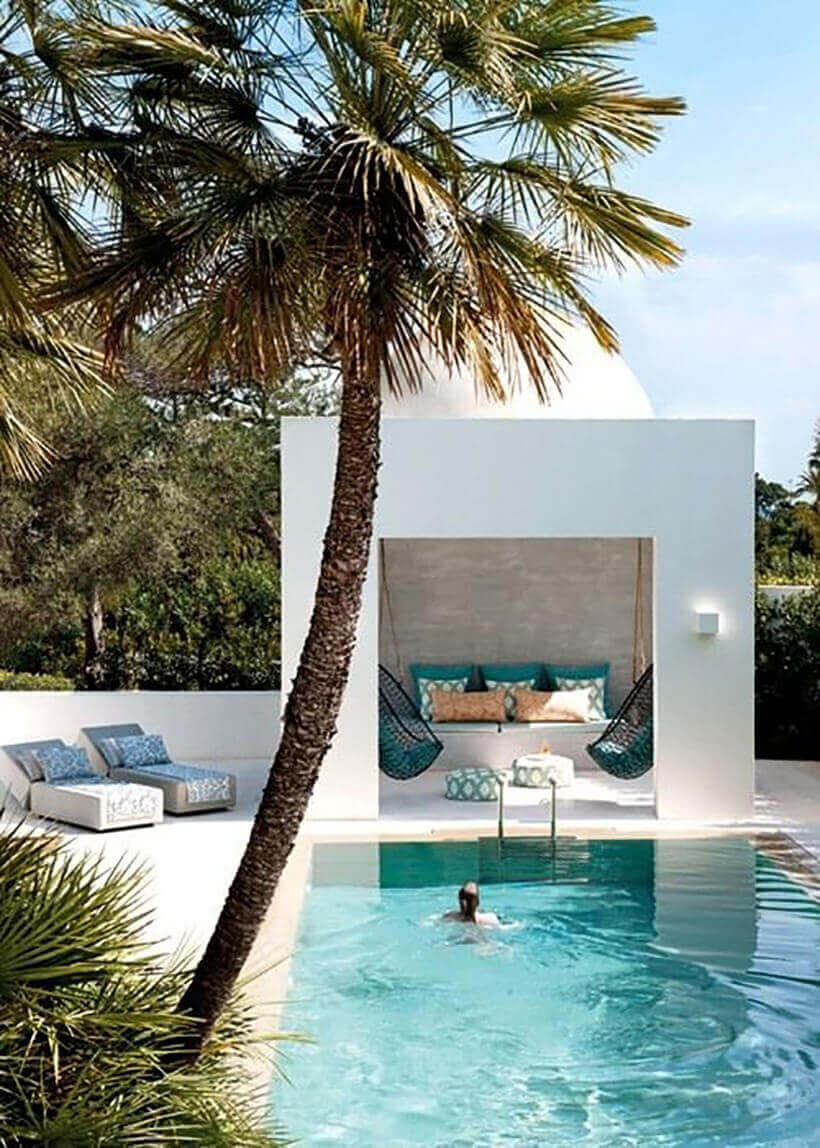 Contemporary swim spa features a rectangular pool with stone decking, modern loungers, and a cabana, surrounded by palm trees.