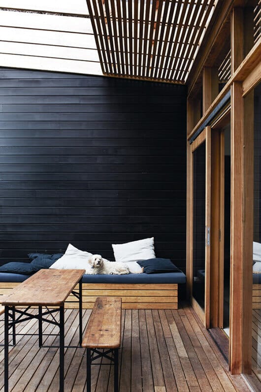 Outdoor seating area with a raised wood deck, featuring a navy blue cushioned bench, rustic timber table, and sun-filtering trellis for shade.