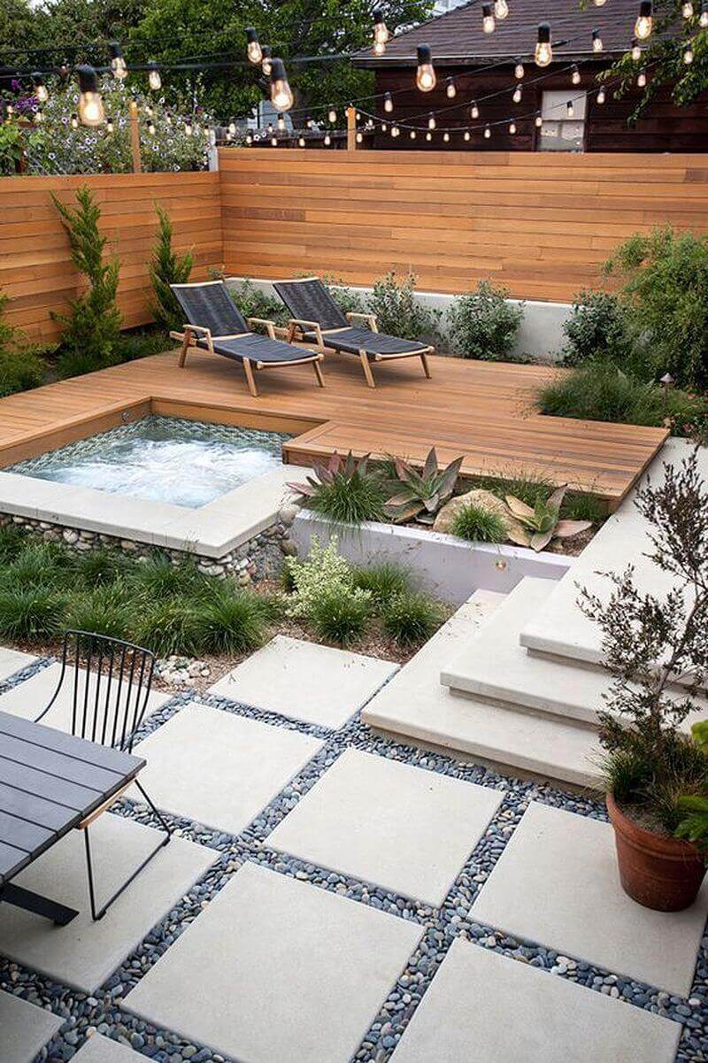 Modern outdoor living space with a wooden deck surrounding a hot tub, featuring reclining chairs and serene landscaping with string lights.