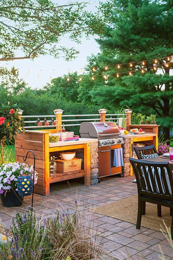 Outdoor kitchen with stone and wood structure beside a rectangular pool. Features a stainless steel grill, countertop, and ambient string lights.