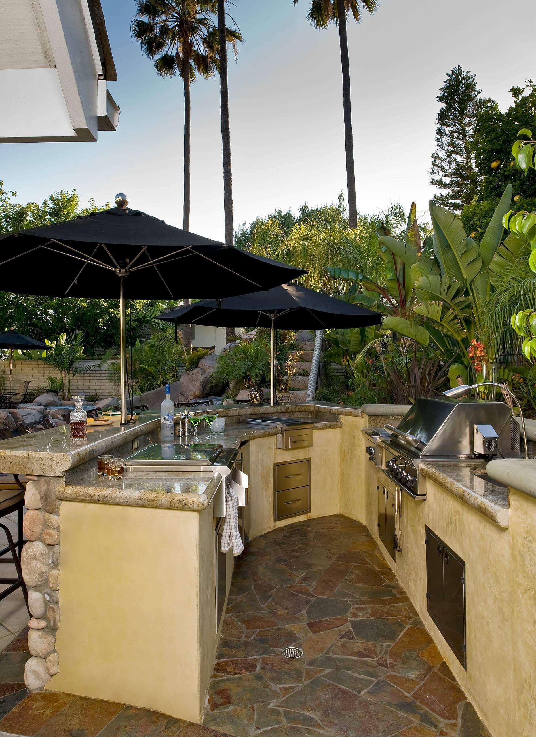 Outdoor kitchen with polished stone countertops, stainless steel appliances, and black umbrellas, surrounded by lush tropical landscaping and a pool.