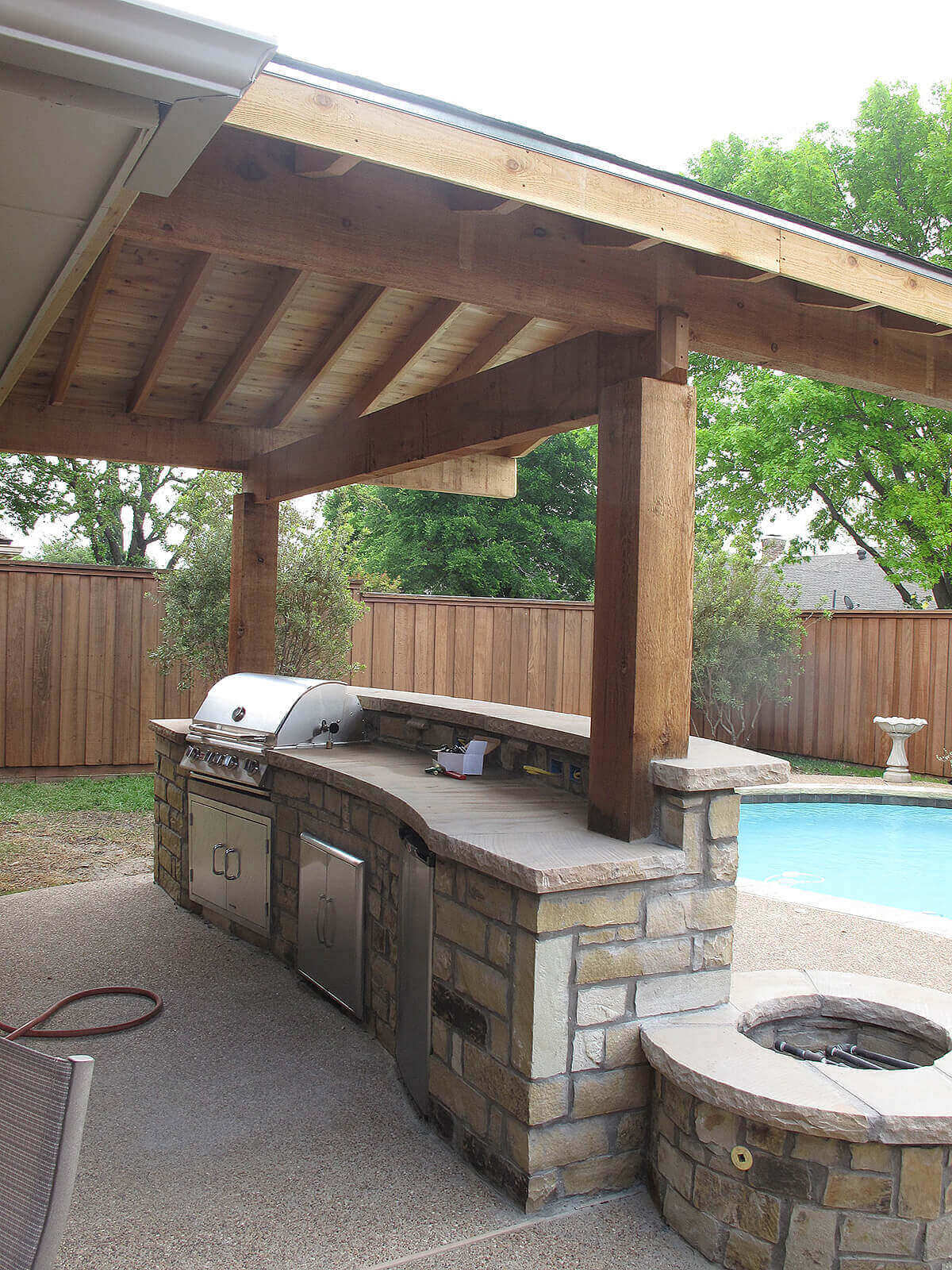 Curved stone countertop outdoor kitchen with stainless steel appliances next to a pool, featuring timber beams and a fire pit for socializing.