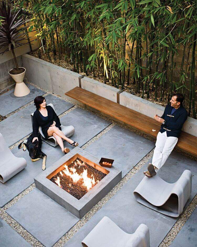 Outdoor fire pit area with a square concrete pit, surrounded by gray pavers and gravel. Features sculptural concrete chairs and a wooden bench, with lush bamboo for privacy. Ideal for cool evenings.