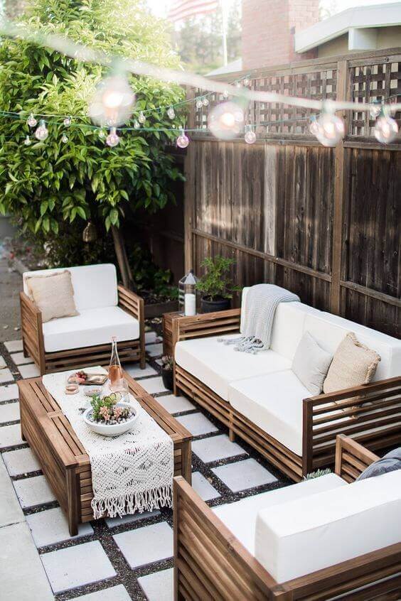 Modern patio featuring modular seating with wooden frames and white cushions, surrounded by a low coffee table, string lights, and lush greenery.
