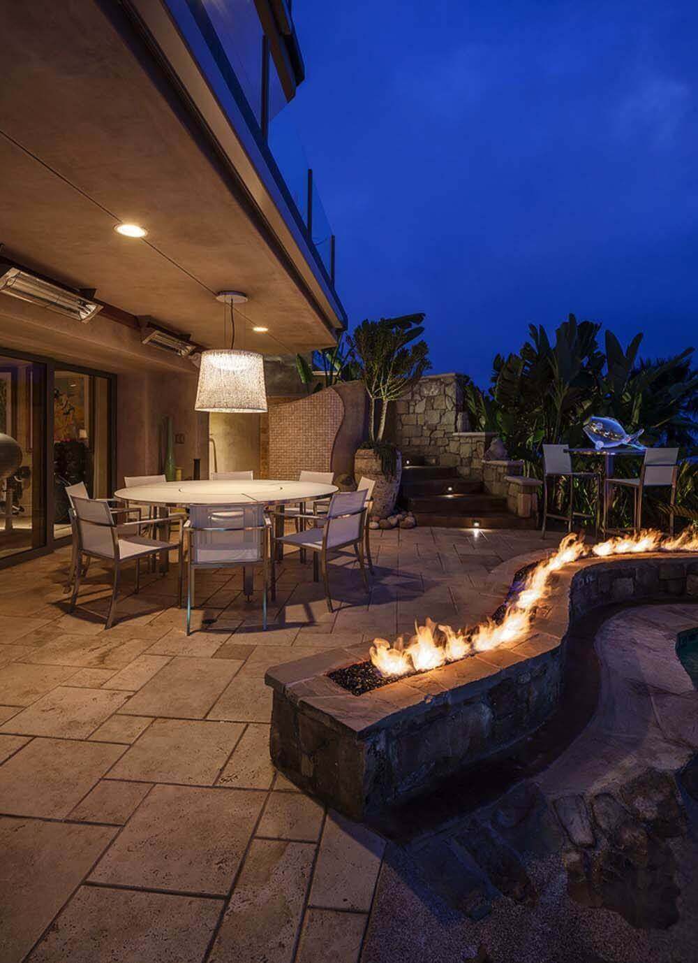 Modern outdoor living area featuring a round table for eight, aluminum chairs with weather-resistant fabric, and a linear stone fire pit.