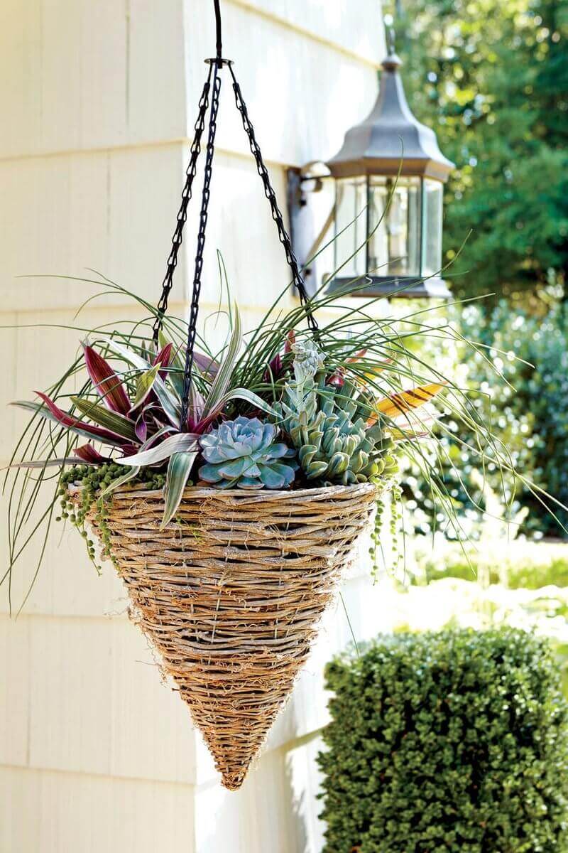 A conical hanging basket made of woven natural materials, featuring succulents and ornamental grasses in green, purple, and silver hues.