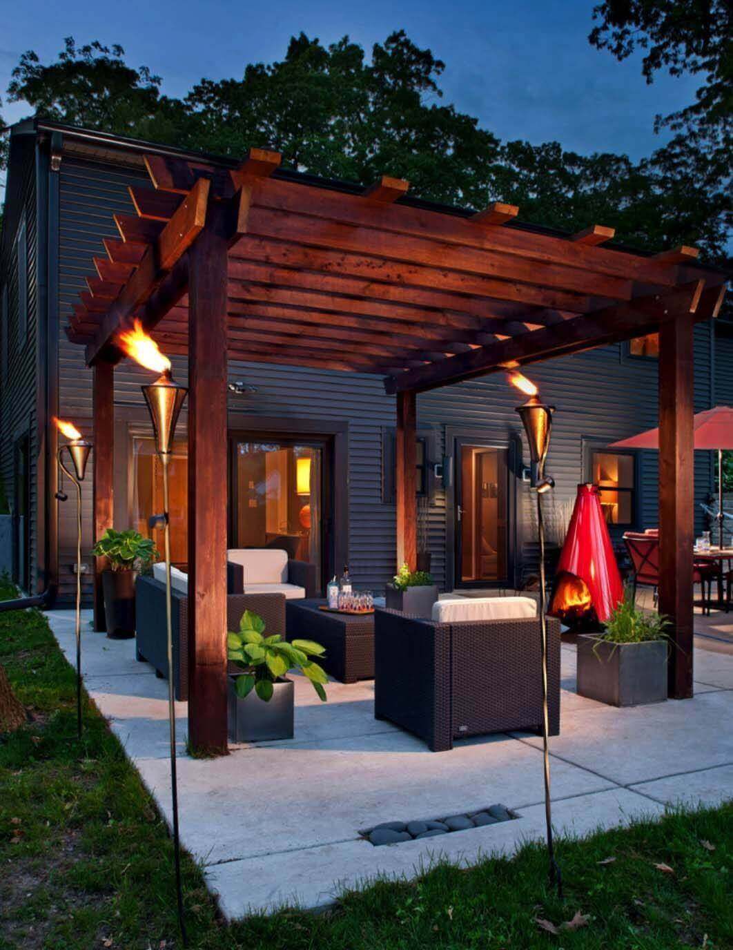 Modern outdoor living space with a dark-stained wooden pergola, concrete patio, contemporary wicker sofas, and lush greenery. Ideal for cozy gatherings.