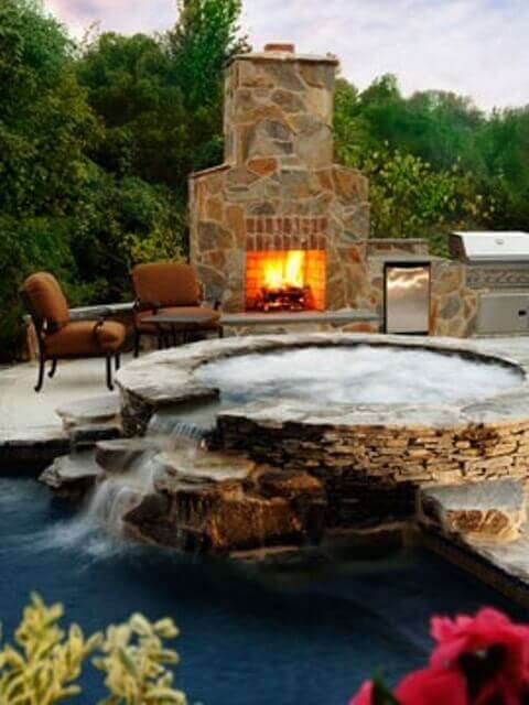 Circular hot tub made of natural stone, surrounded by a flagstone patio, seating area, and a natural stone fireplace. Peaceful backyard spa.