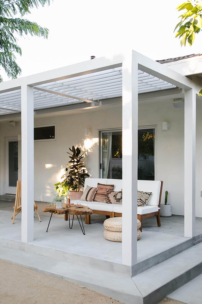 Modern patio featuring a concrete floor, white pergola, minimalist couch, woven pouf, rustic coffee table, and terracotta plants.