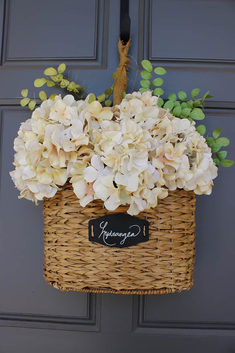Artificial hydrangea blooms in beige and white, with green eucalyptus, arranged in a woven wicker basket for outdoor decor.