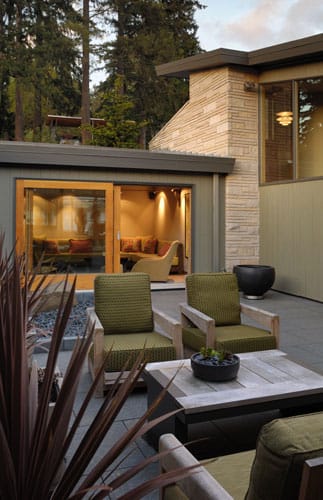 Modern outdoor living space with two wooden lounge chairs, olive green cushions, and a low coffee table on slate tile flooring, surrounded by coniferous trees.