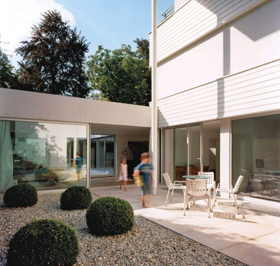 Modern outdoor space featuring low-maintenance gravel, rounded boxwood shrubs, a spacious patio with light stone slabs, and a round table with lightweight chairs.