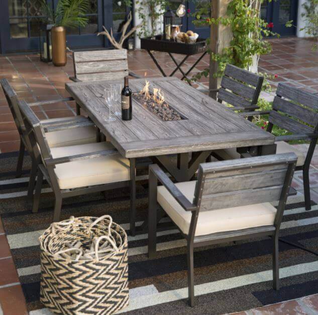 Rectangular wooden dining table with a central fire pit, six cushioned chairs, outdoor rug, and woven basket, set for gatherings.