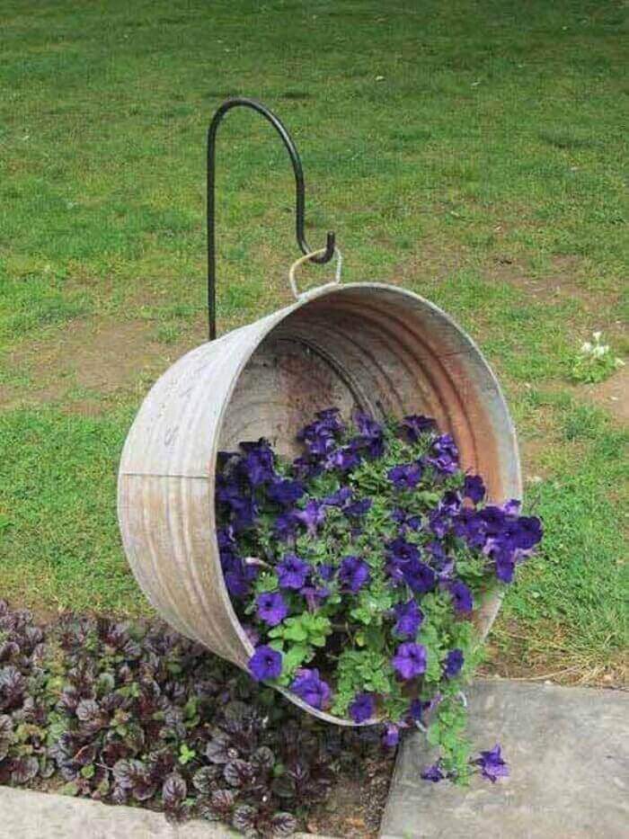 Rusty metal bucket hanging planter filled with vibrant purple petunias, surrounded by dark burgundy foliage, set in a temperate garden.
