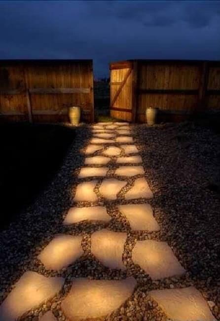 Illuminated pathway with irregular stone slabs on gravel, framed by wooden gates and fencing, creating a rustic, inviting entrance.
