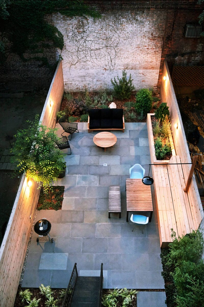 Patio area with stone pavers, featuring a black sofa, wooden chairs, and a round coffee table. Surrounded by greenery and a grill.