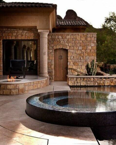 Circular hot tub with a polished black stone surface, surrounded by light-colored flagstone patio, rustic stone wall, and modern fire pit.