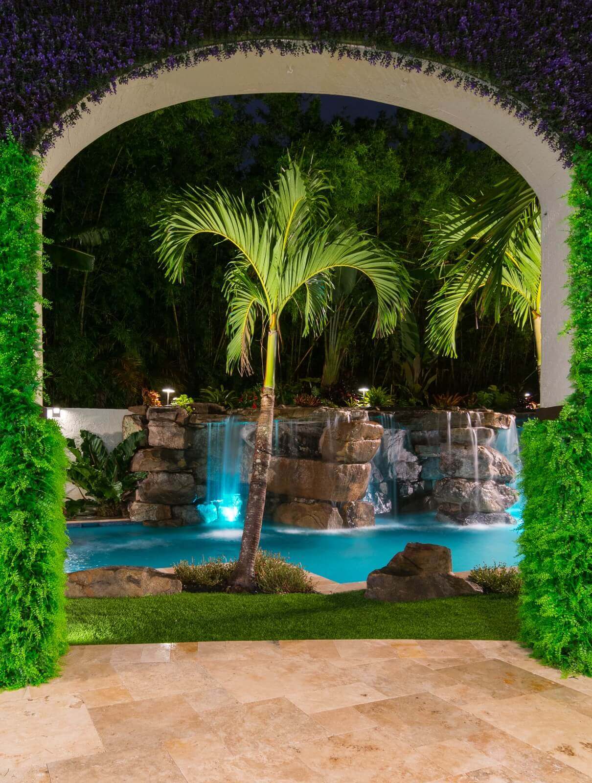 Vibrant tropical oasis featuring a rectangular pool surrounded by lush greenery, palm trees, and a cascading stone waterfall.
