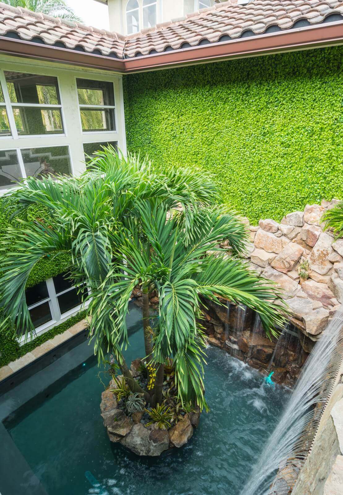 Rectangular pool featuring a cascading waterfall and a central palm tree, surrounded by lush greenery and rocky landscaping, creating a tropical oasis.