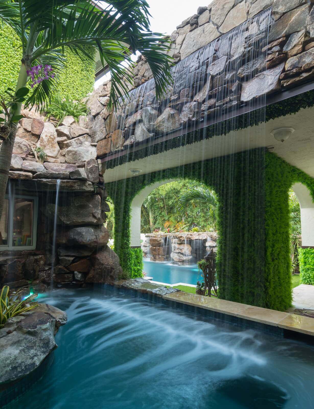 Cascading waterfall with natural stone, flowing into a shallow pool surrounded by tropical plants, enhancing a serene outdoor space.