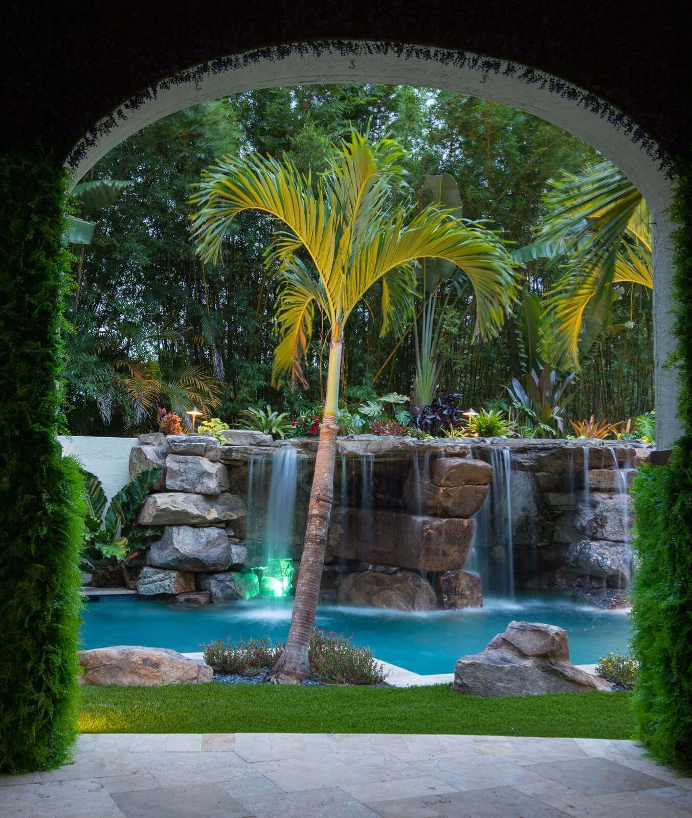 A cascading waterfall flows into a turquoise pool, surrounded by tropical plants and boulders, creating a serene backyard oasis.