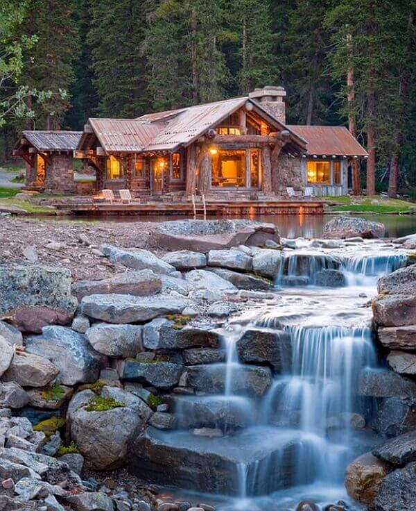 A rustic cabin with a weathered metal roof beside a tranquil pond, featuring a custom rectangle pool and a stone waterfall.