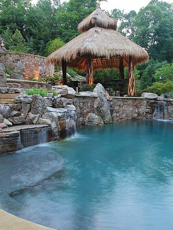 Naturalistic pool with waterfalls and stone formations, surrounded by boulders and a thatched pavilion for shaded seating.