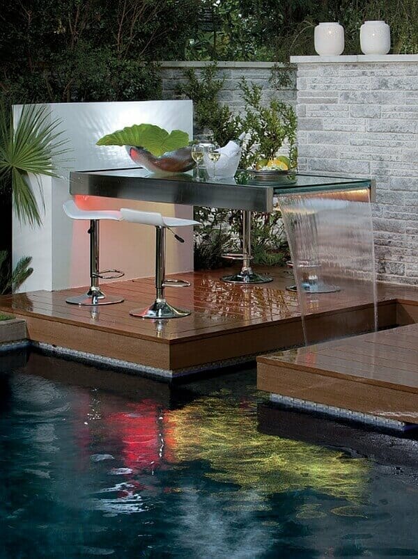 Outdoor bar with a sleek stainless steel counter and glass top, overlooking a pool with a gentle water feature, surrounded by tropical plants.