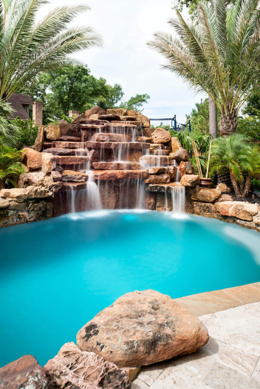 A serene swimming pool features a natural stone waterfall, framed by tan coping and surrounded by lush greenery, creating a tropical oasis.