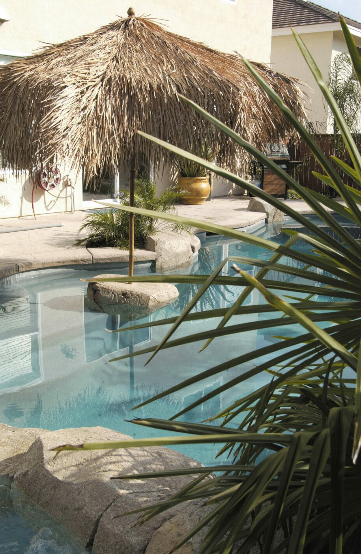 Lush pool area with a thatched umbrella, smooth boulder, tropical plants, and textured concrete decking for relaxation. Ideal for custom pools.