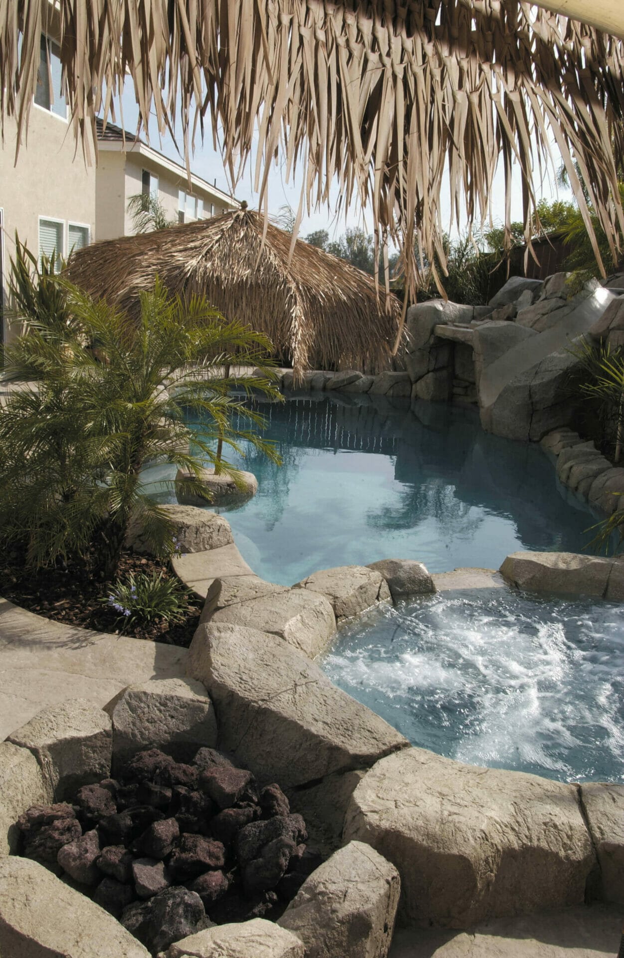 Custom swimming pool with naturalistic rock formations and waterfalls, surrounded by palm trees and a fire pit, creating a tropical oasis.
