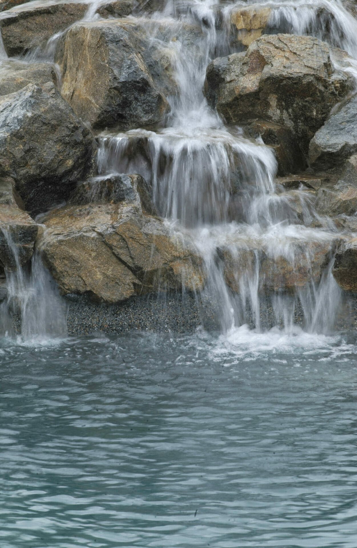Waterfall cascades over granite boulders into a shallow pool, creating a serene focal point in a minimalist landscape.