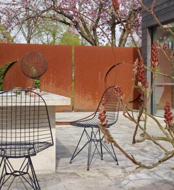 Contemporary outdoor dining area featuring a honed concrete table and minimalist black wireframe chairs, surrounded by lush greenery and corten steel panels.