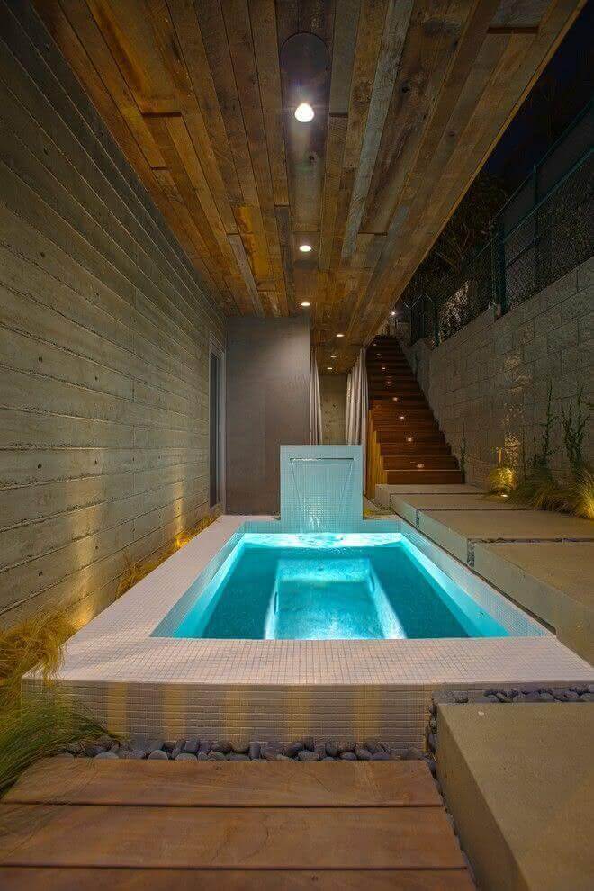 Rectangular spa with white mosaic tile, under a wooden overhang, surrounded by concrete steps and native grasses, creating a serene atmosphere.
